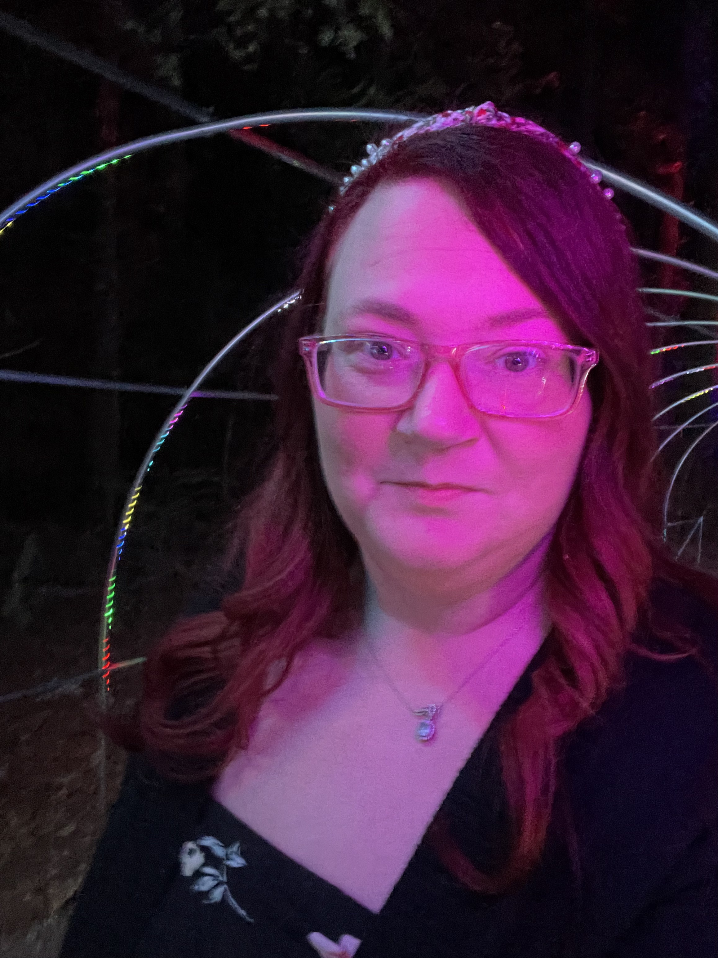 Ashley A. Love poses for a close-up selfie under vivid purple and pink event lighting, wearing glasses and a black top.