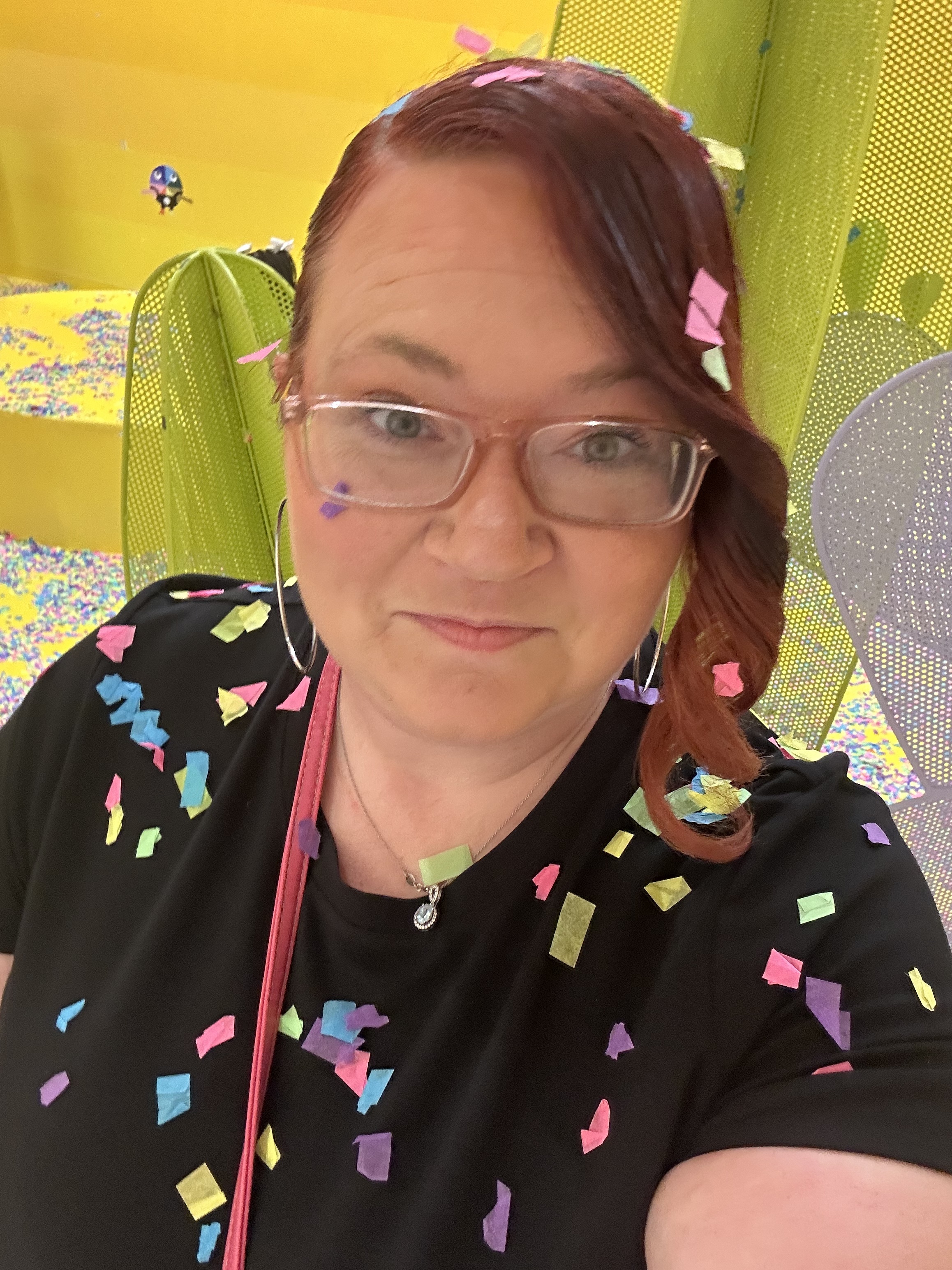 Ashley A. Love smiles for a close-up selfie in a colorful indoor play space, wearing glasses and a black top sprinkled with bright paper confetti.