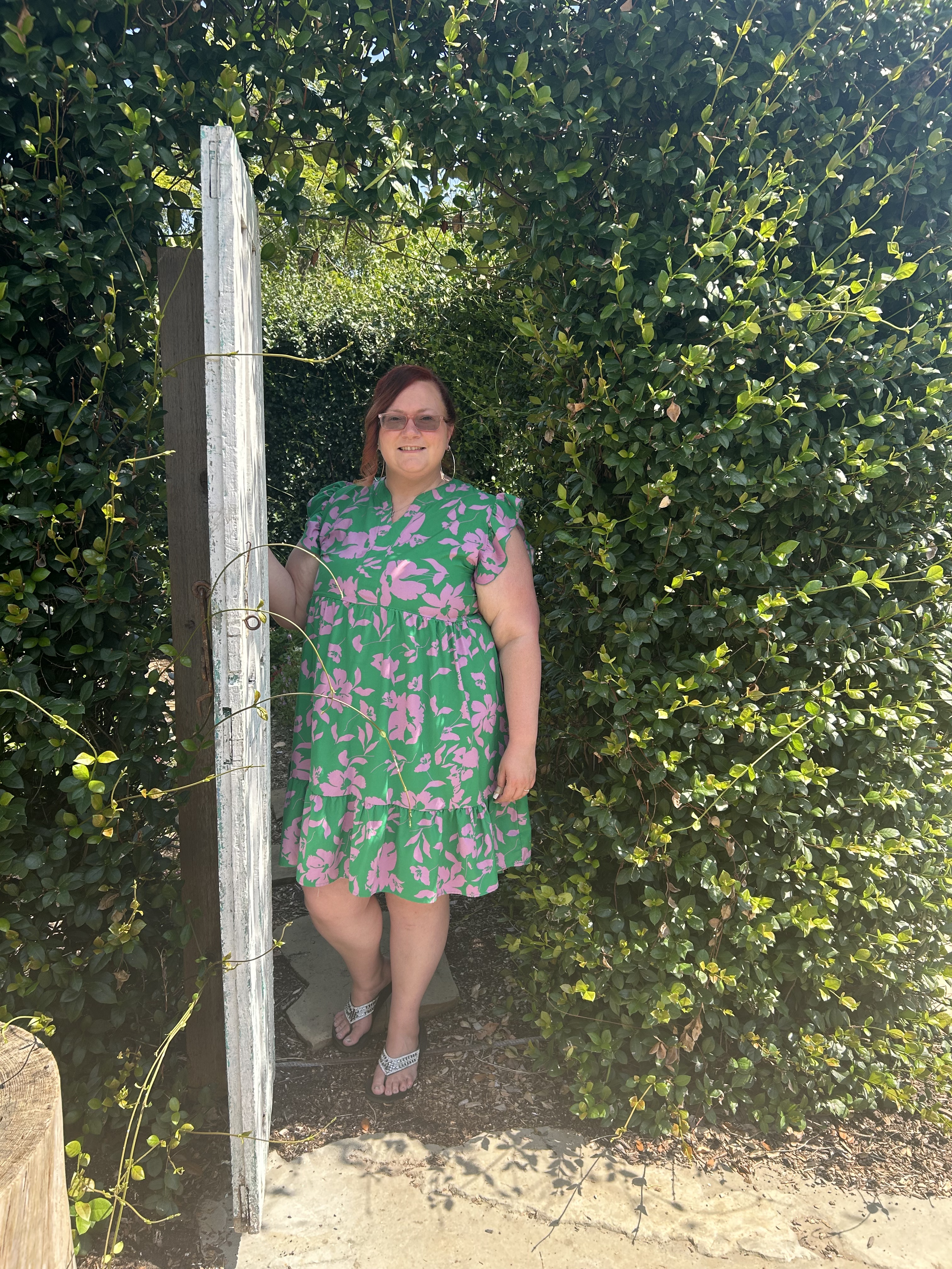 Ashley A. Love stands in a green floral dress beside a weathered garden gate, surrounded by sunlit hedges and climbing greenery.